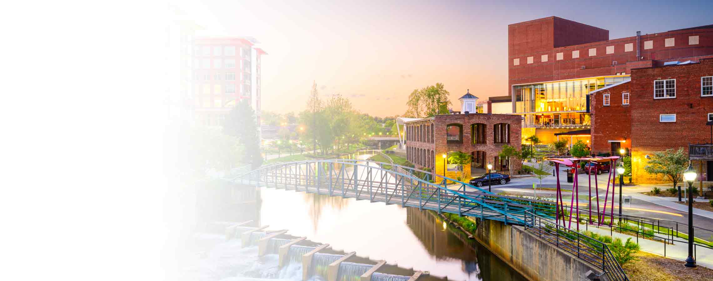 A sunset photo showing a walking bridge over a downtown Greenville river.